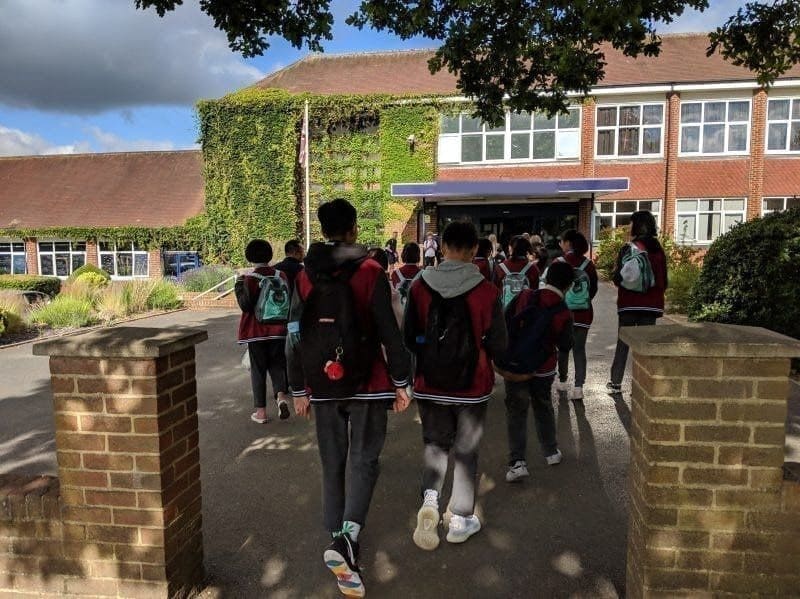 Students with rucksacks, seen from behind, walking towards the entrance of a partner school
