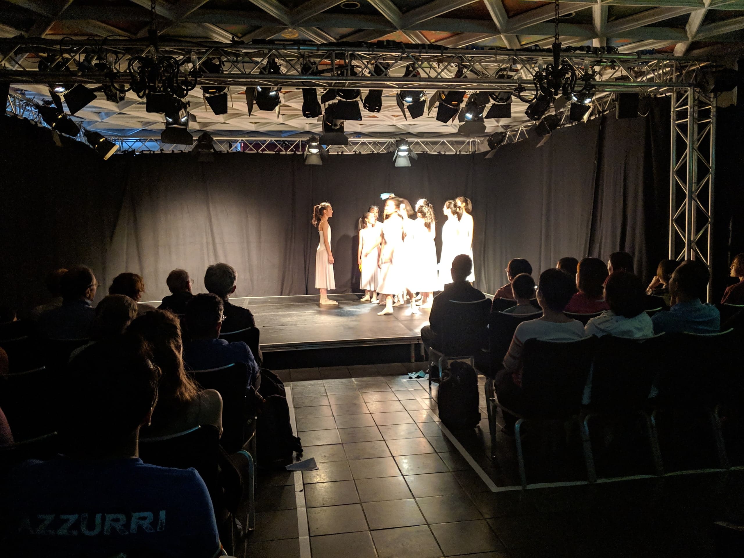 Dancers in white performing The Moment under stage lights, with the audience in silhouette